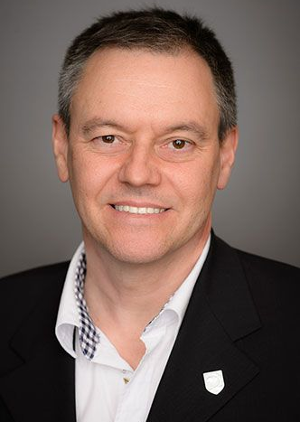 Un homme en blazer sourit, vêtu d'une chemise à motifs. Fond gris.