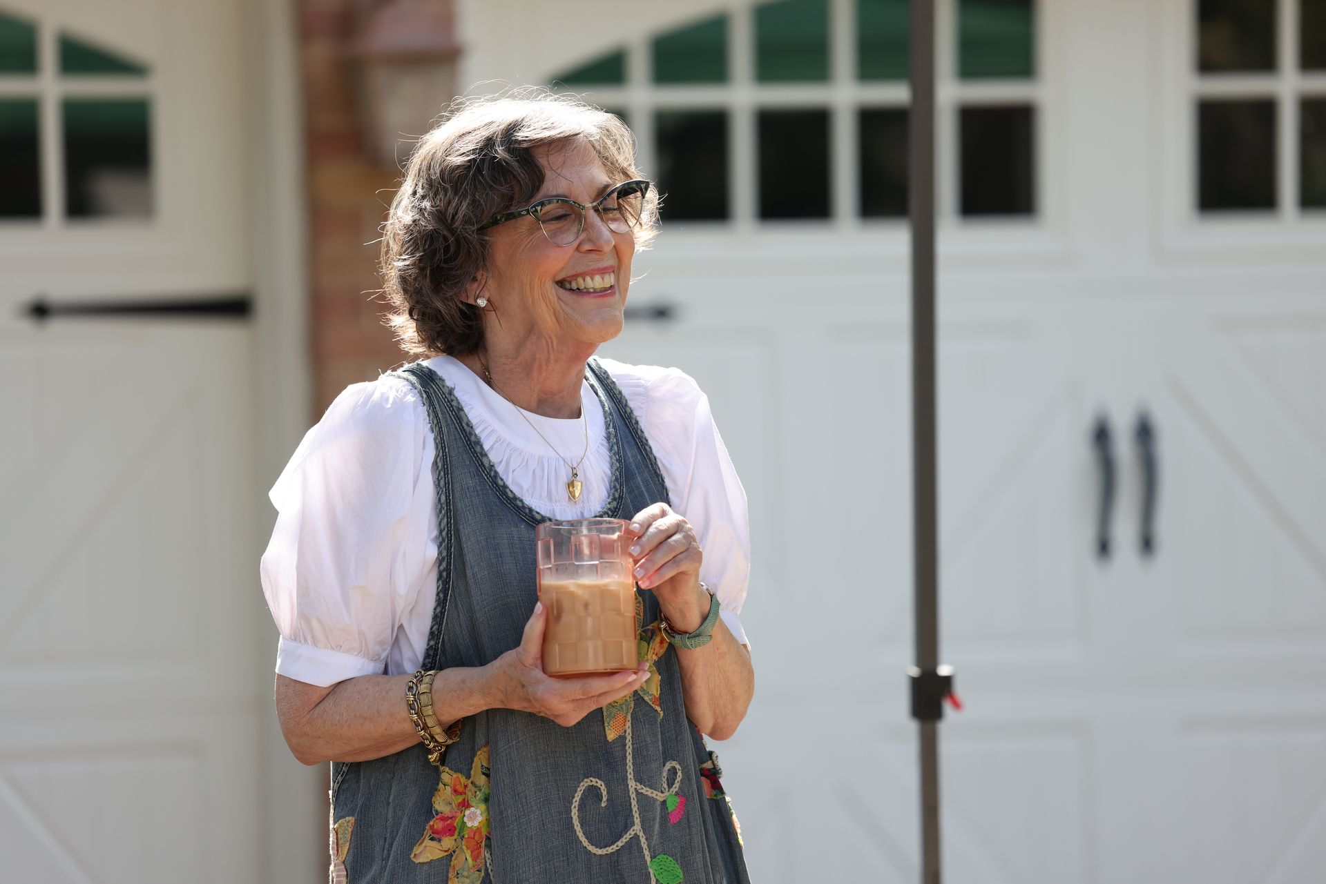 An elderly woman is holding a glass of milk and smiling in front of a garage door.