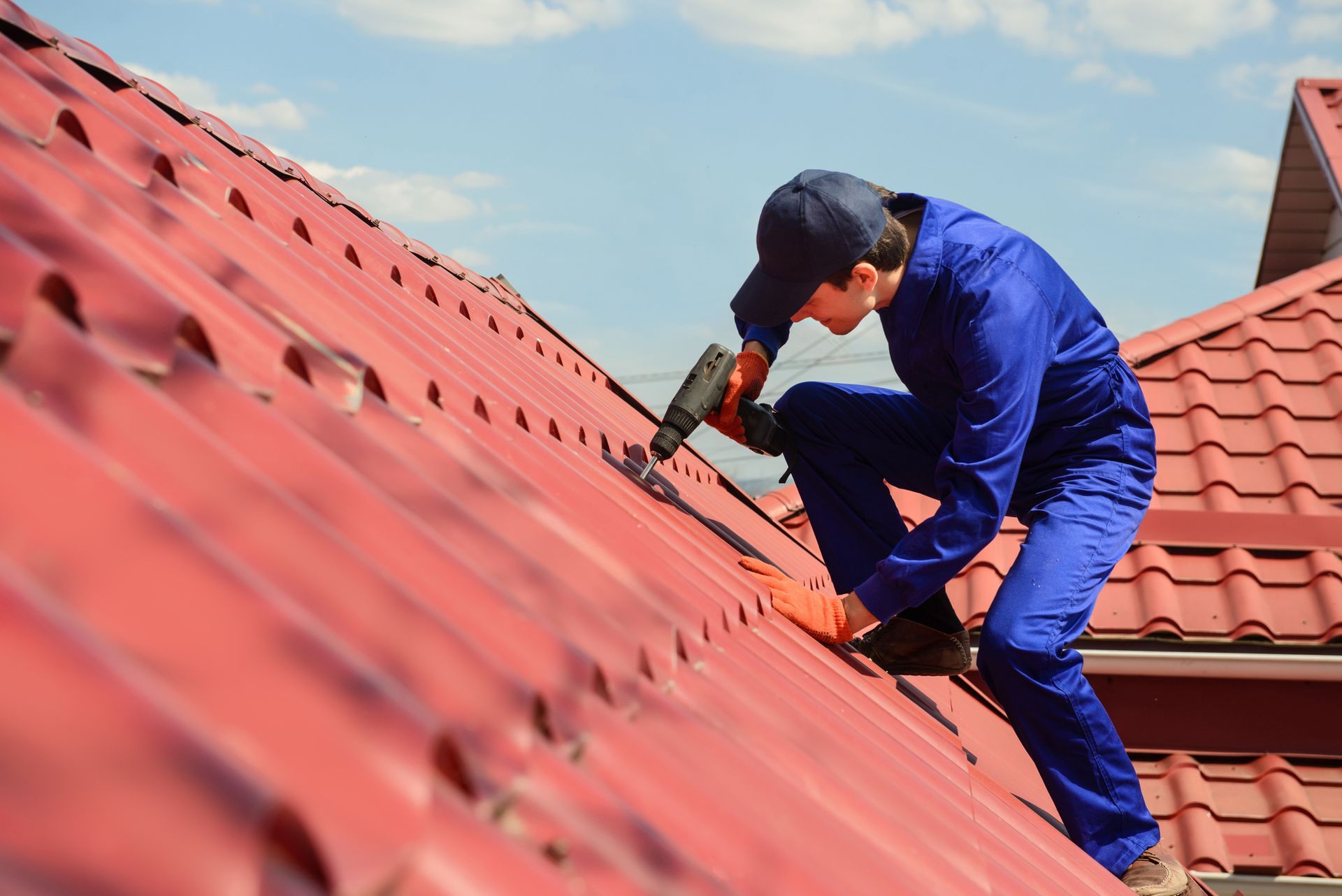 A male roof contractor in a blue uniform is working with a power drill on a red roof in daylight.