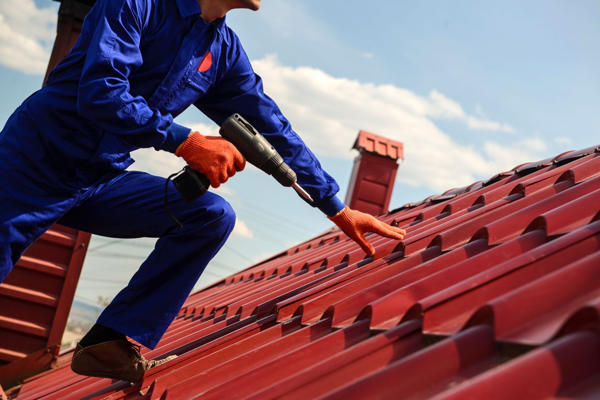Roof contractor installing with drill in hand red metal panels on residential roof under blue sky.