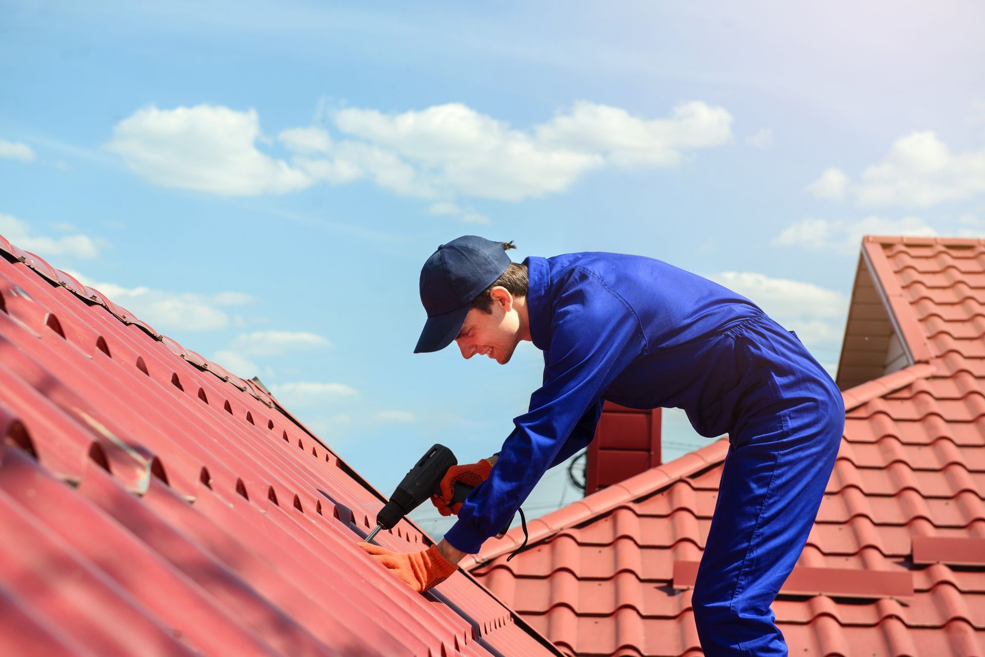 Roof contractor installing with drill in hand red metal panels on residential roof under blue sky.
