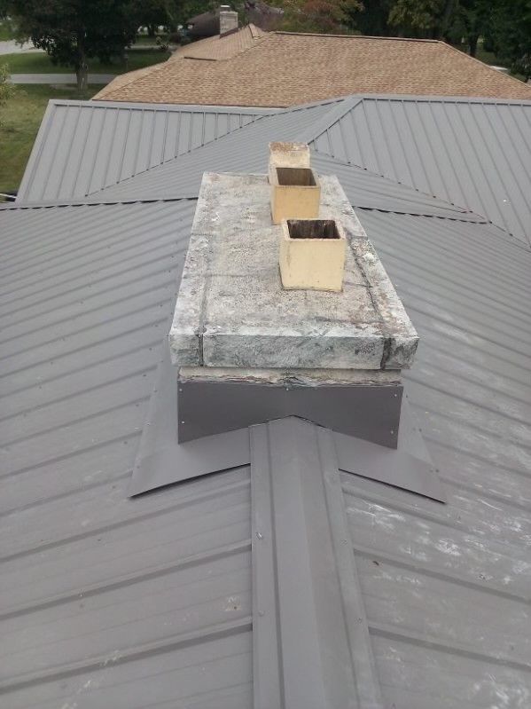 Chimney on a gray metal roof, with two tan flues. The chimney is made of weathered concrete.
