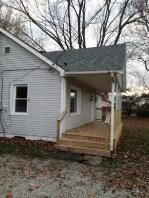 Side view of a light gray house with a wooden porch and stairs. The porch has a roof and is partially enclosed.