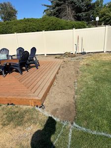 A wooden deck with chairs and a table in a backyard with a white fence.