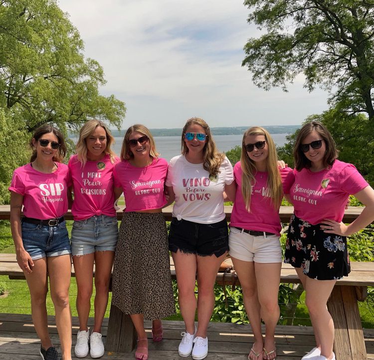Girls wearing matching pink shirts, and one girl wearing white. At a winery in front of a lake. 