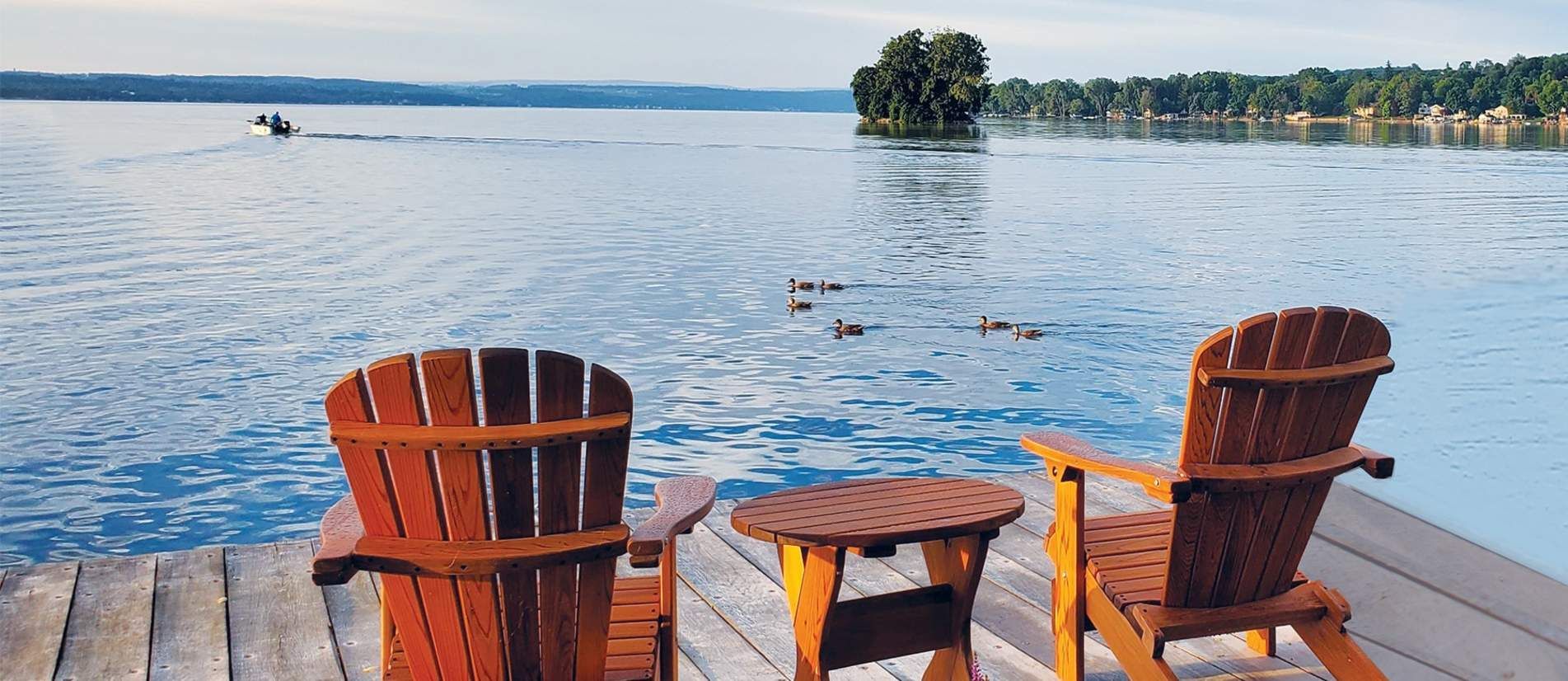 Two redish brown Adirondack chairs facing out in front of a gorgeous lake on a wooden dock