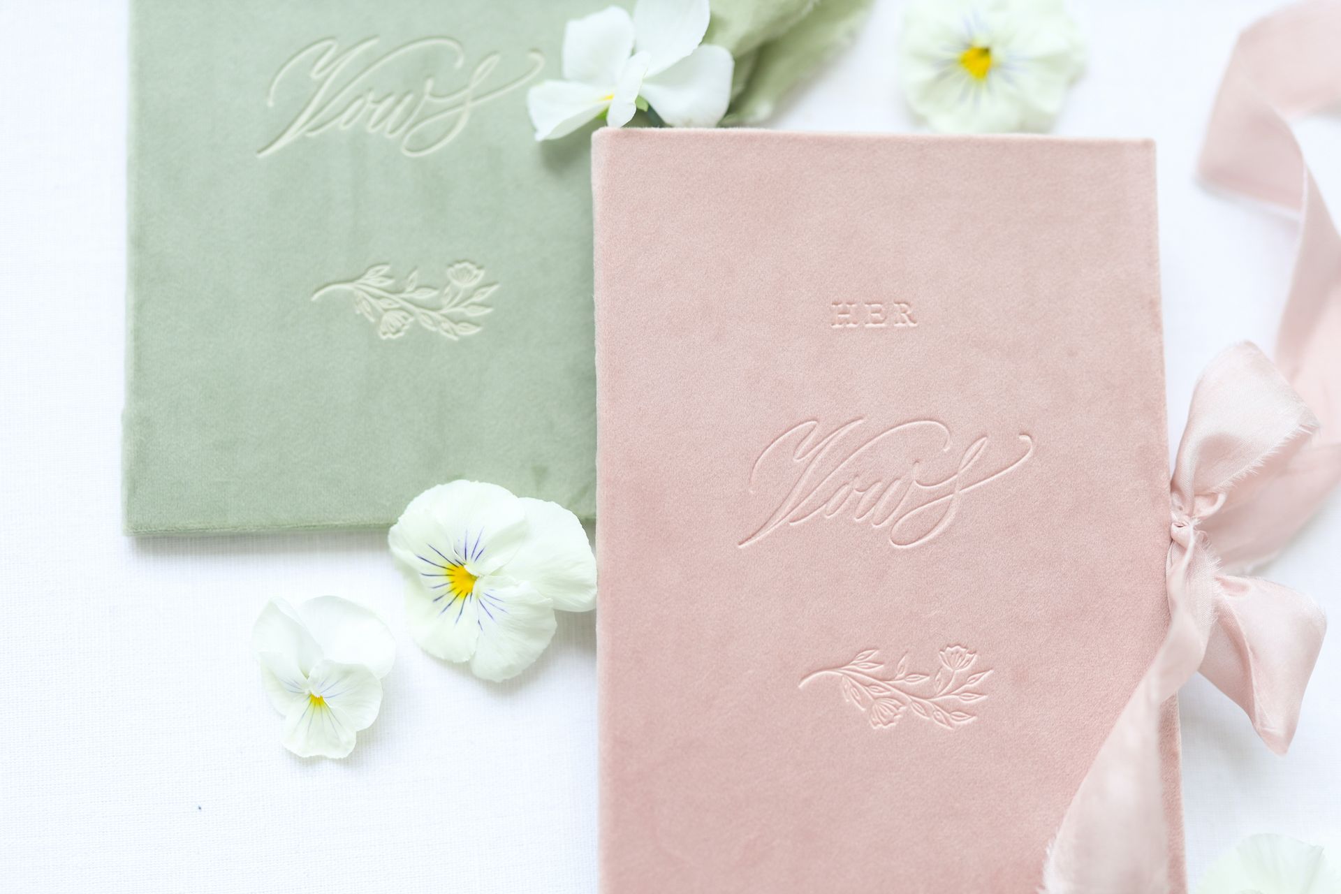 Two books for writing vows - one in pink and one in green, laid on top of a while table with flowers