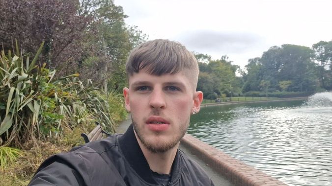 Man standing near a lake with a fountain. He's wearing a black jacket, in an outdoor park setting.
