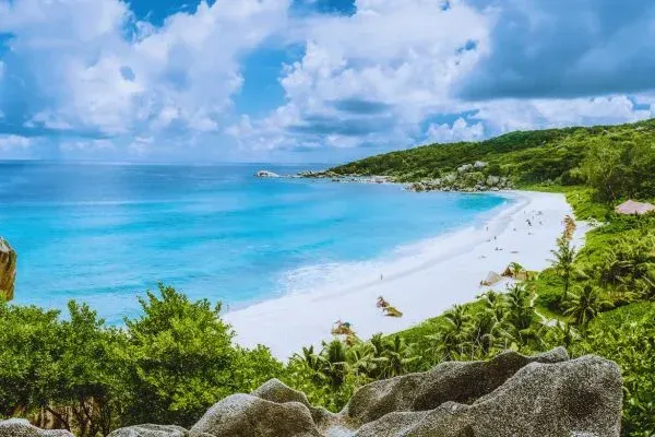 A tropical beach with turquoise water, white sand, green vegetation, and blue sky with clouds.