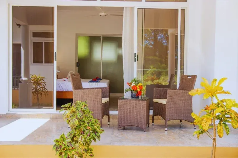 Patio with wicker furniture and sliding doors leading to a bedroom. Sunlight streams in; plants add color.