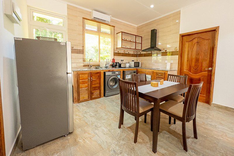 A kitchen with wooden cabinets, a dining table, and a refrigerator. Sunlight streams through a large window.