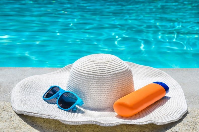 A hat , sunglasses , and sunscreen are sitting on the edge of a swimming pool.