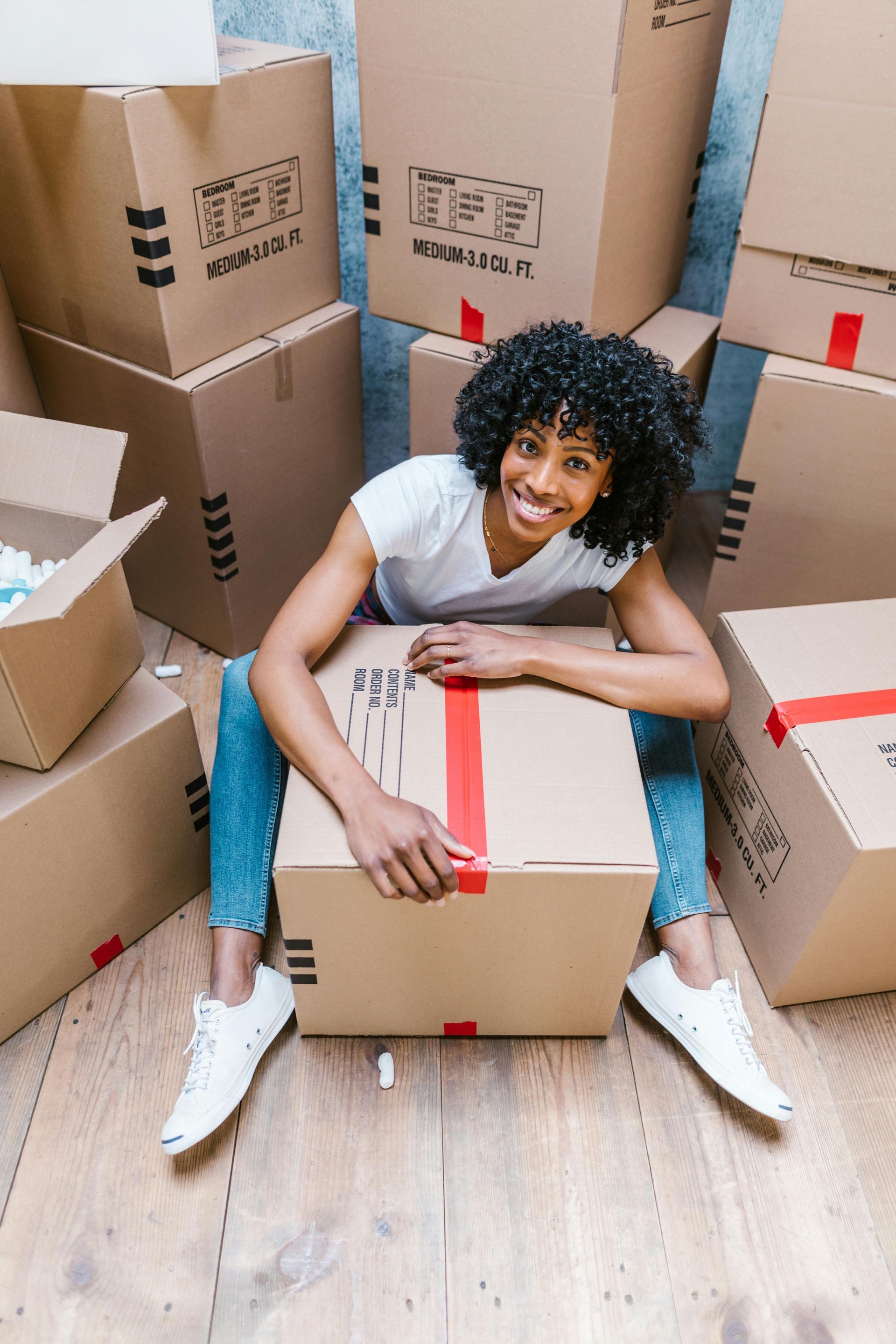 A woman is sitting on a cardboard box surrounded by moving boxes.