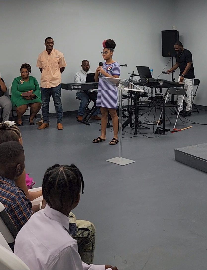 Girl speaking into a microphone, group of people in the background. Inside a room.