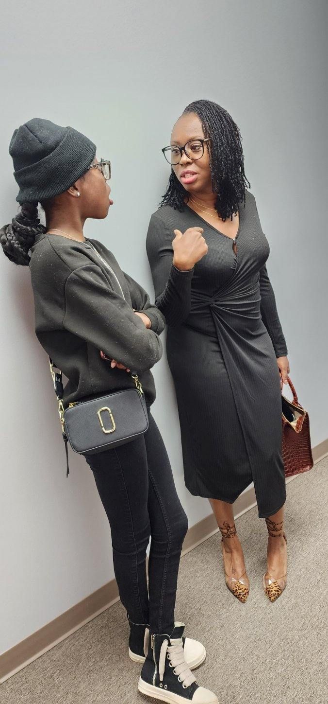 Two women in casual outfits leaning against a wall, talking. One wears a black dress, the other a black hat and sweatshirt.
