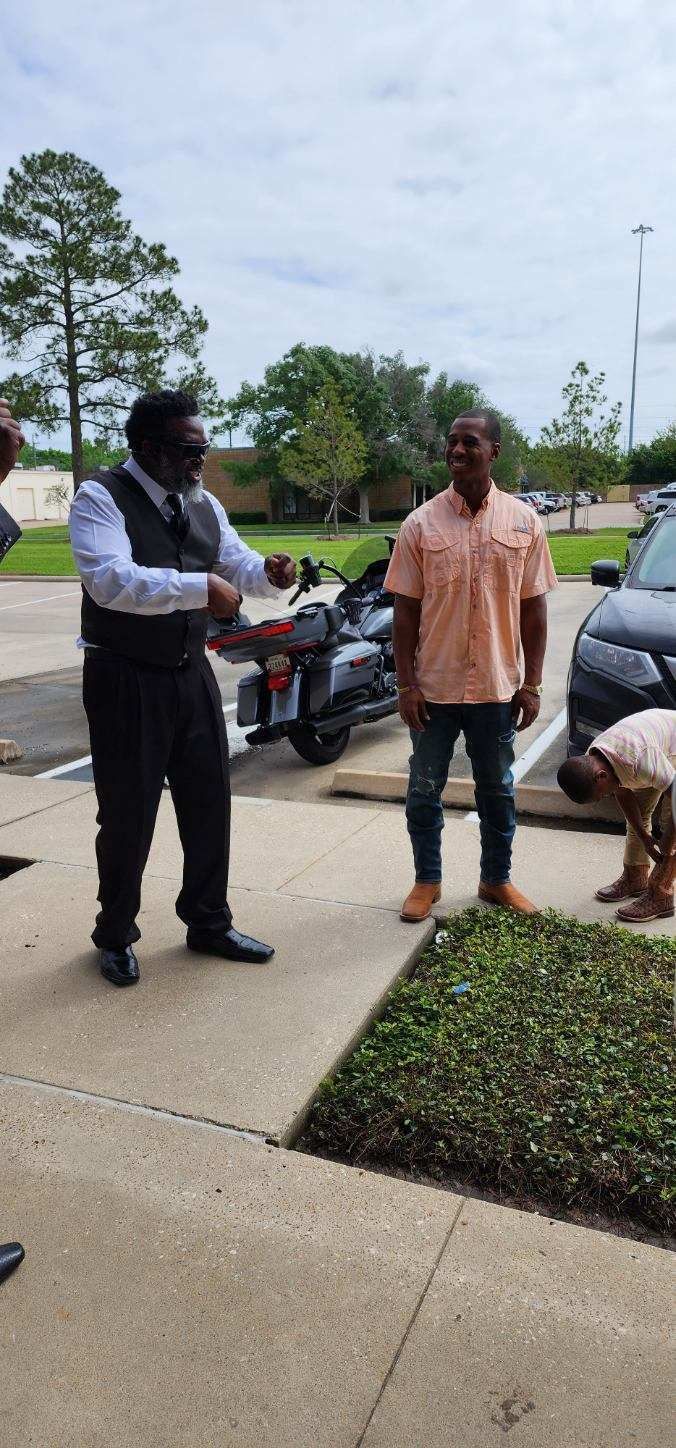 A man in a suit gestures near a motorcycle. Other men are nearby outdoors. Overcast sky.