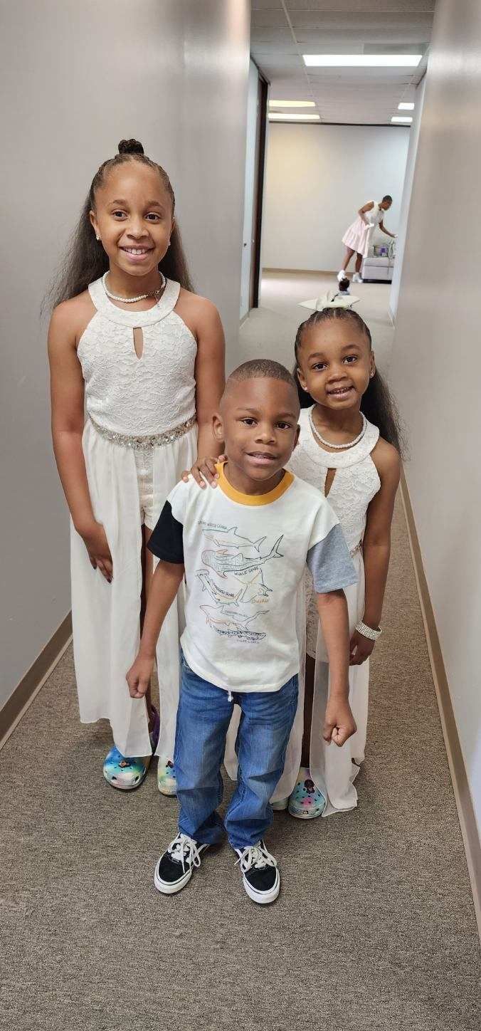 Three children in a hallway: two girls in white dresses smile next to a boy in a t-shirt and jeans.