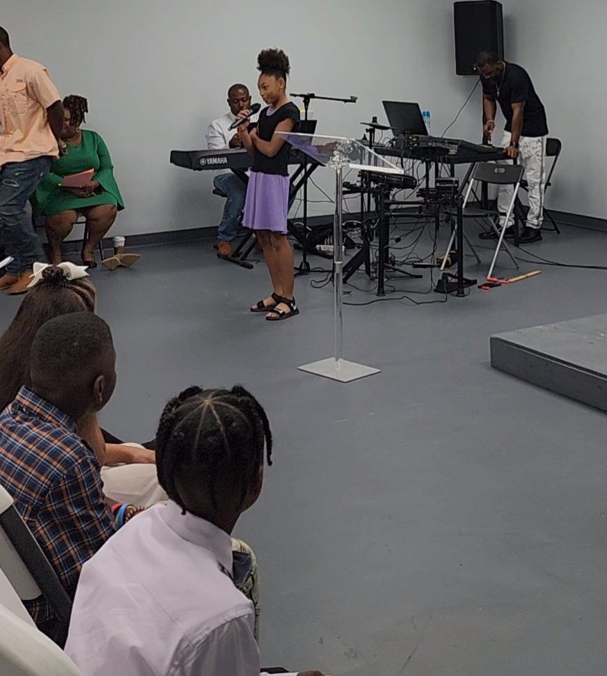 Girl speaking at a podium, in front of a keyboard and audience.