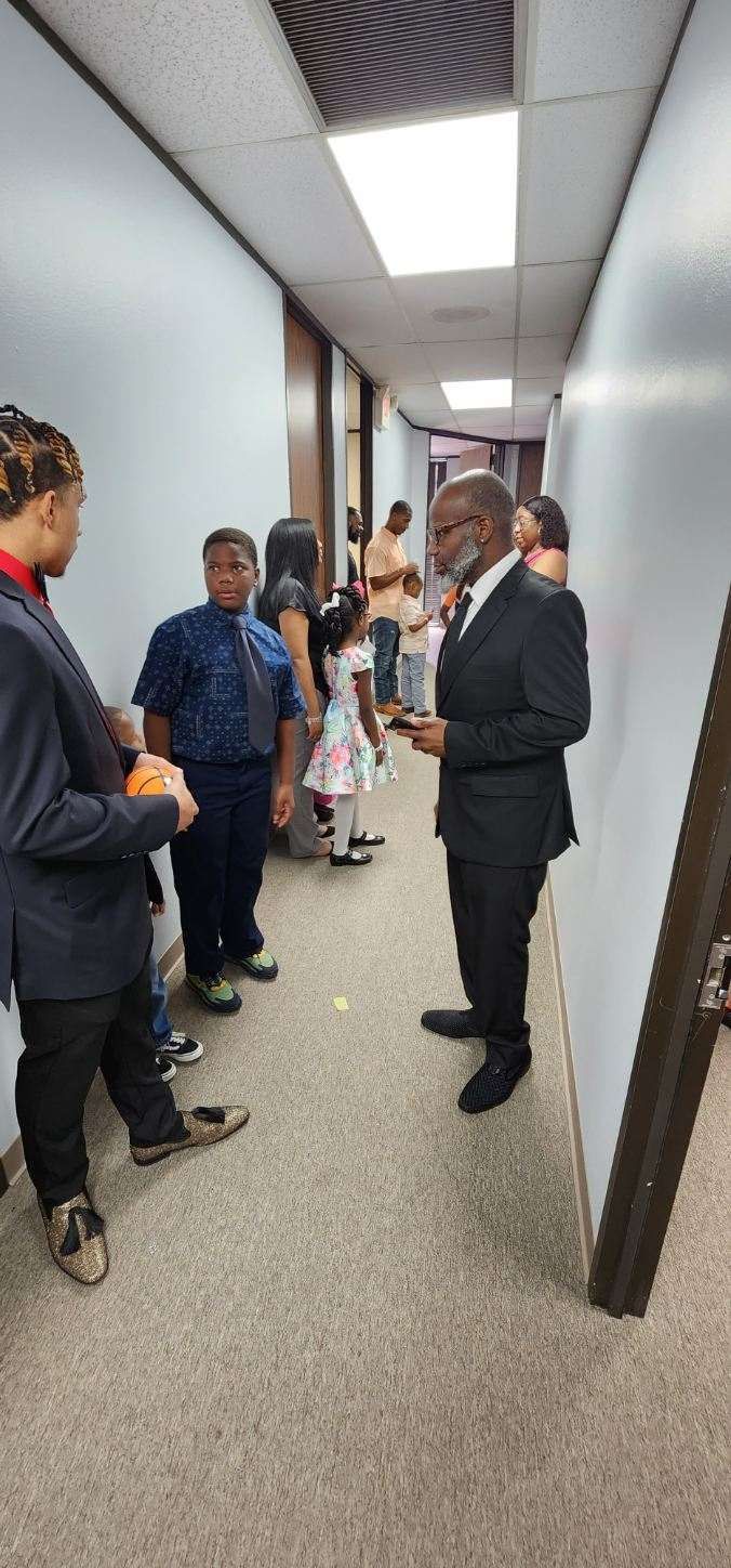 People standing in a hallway, one man in a suit holding a phone. Neutral colors.