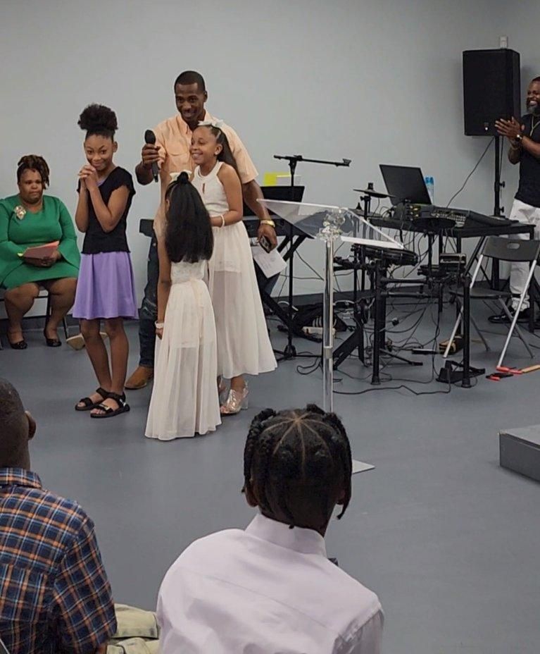 Man with a microphone, two young girls in white dresses, other people clapping, music equipment in a church.