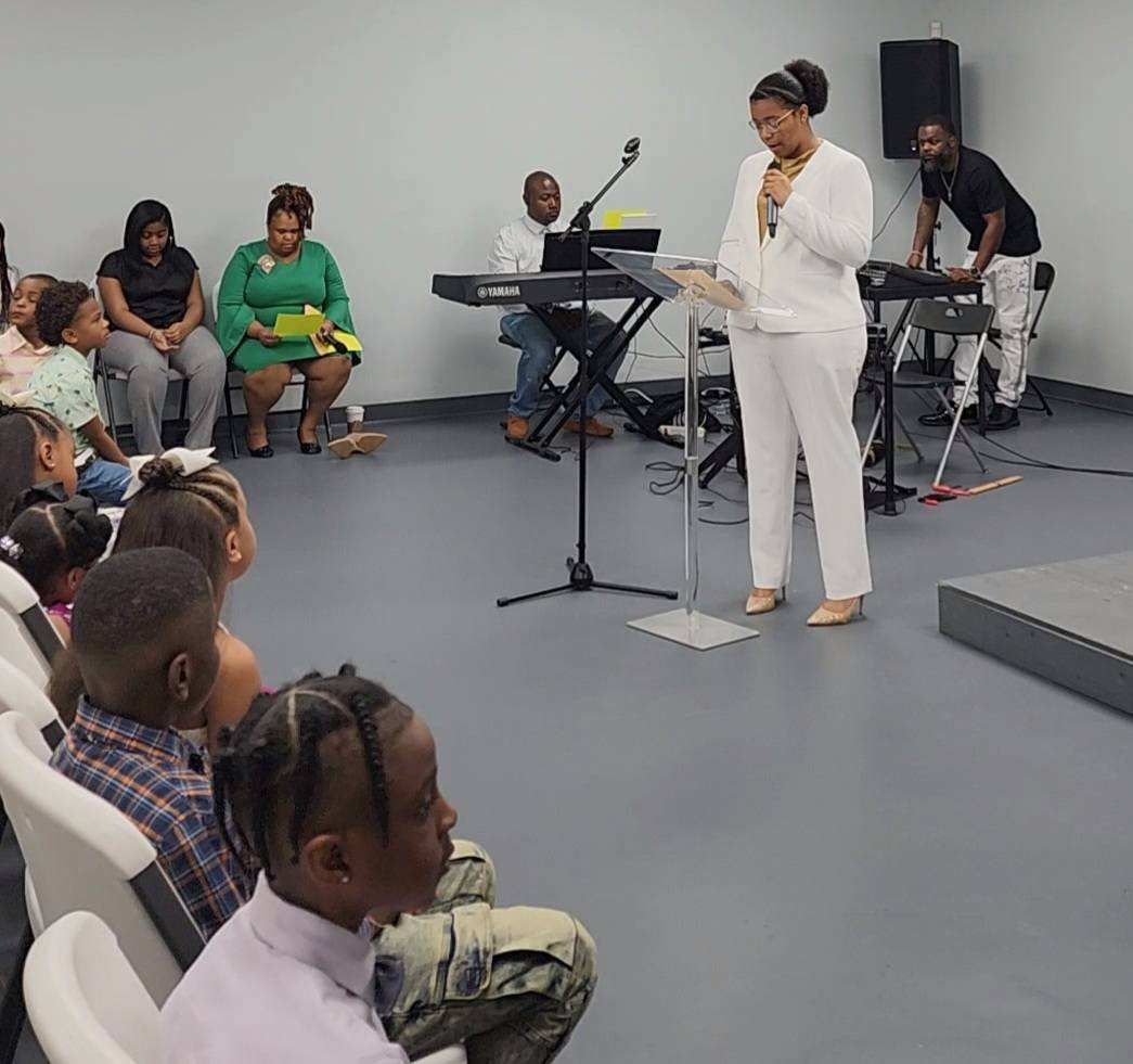 Woman speaking at podium in front of seated children and musicians in a bright room.
