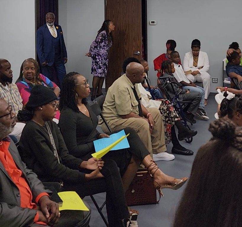 Group of people seated in a room, engaged in a meeting. Some hold papers, dressed in various attire.