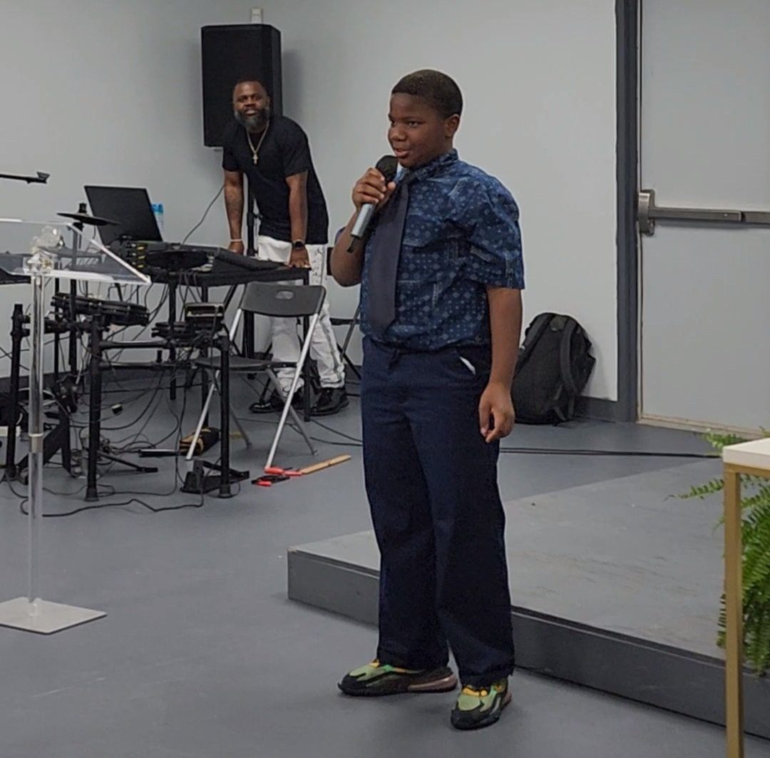 A young boy with a microphone speaks on a stage; a man smiles behind the sound equipment.
