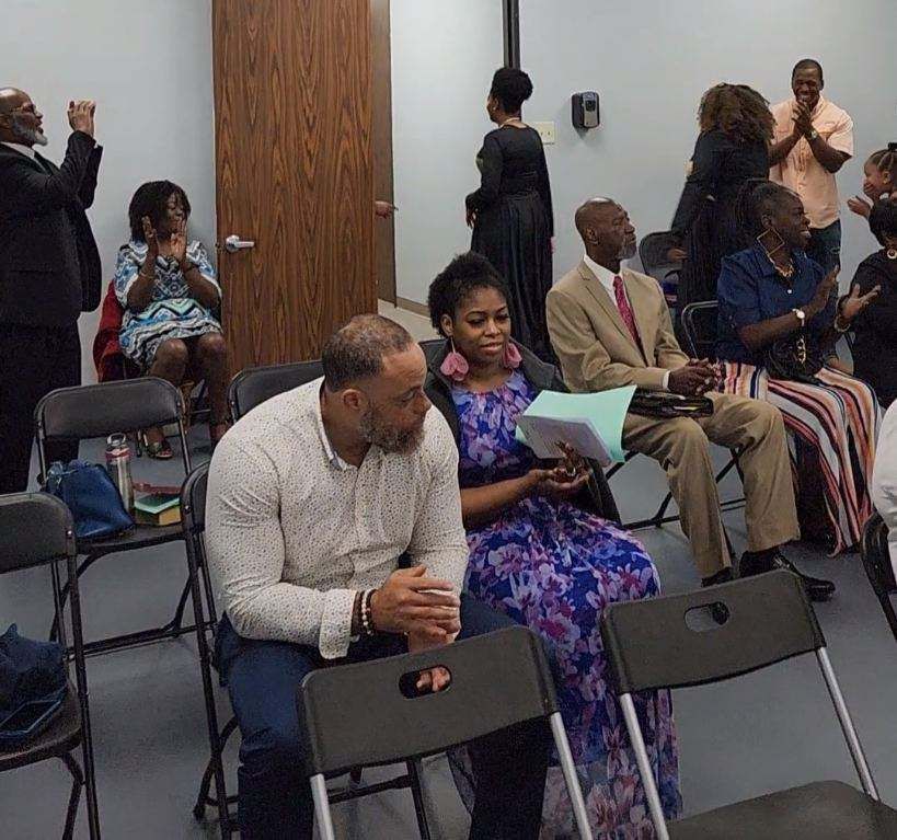 People gathered in a room, clapping. Some sit and others stand. A man and woman are in the front.