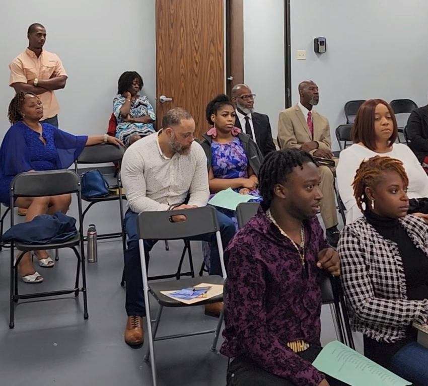 A group of people, primarily Black, seated in a meeting room. Some are listening attentively.