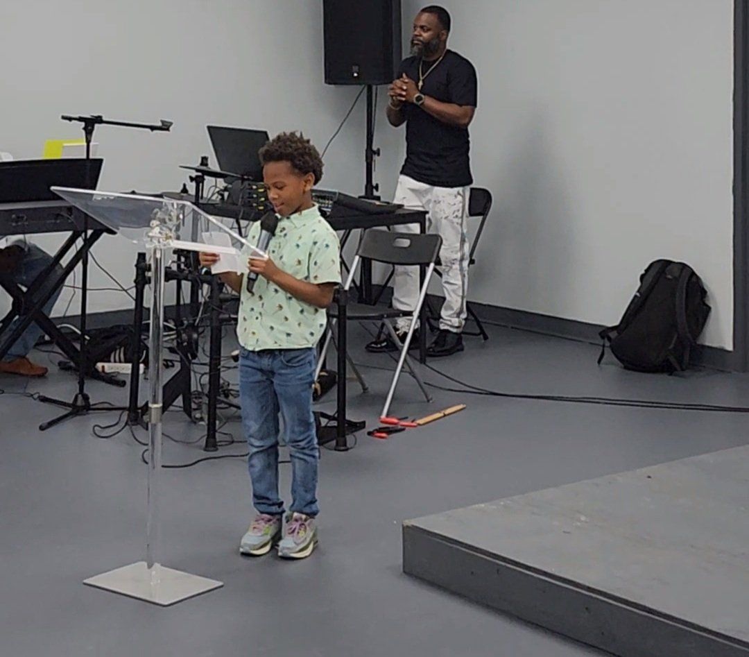 Young boy reading at a podium, adult man in background, musical equipment, modern interior.