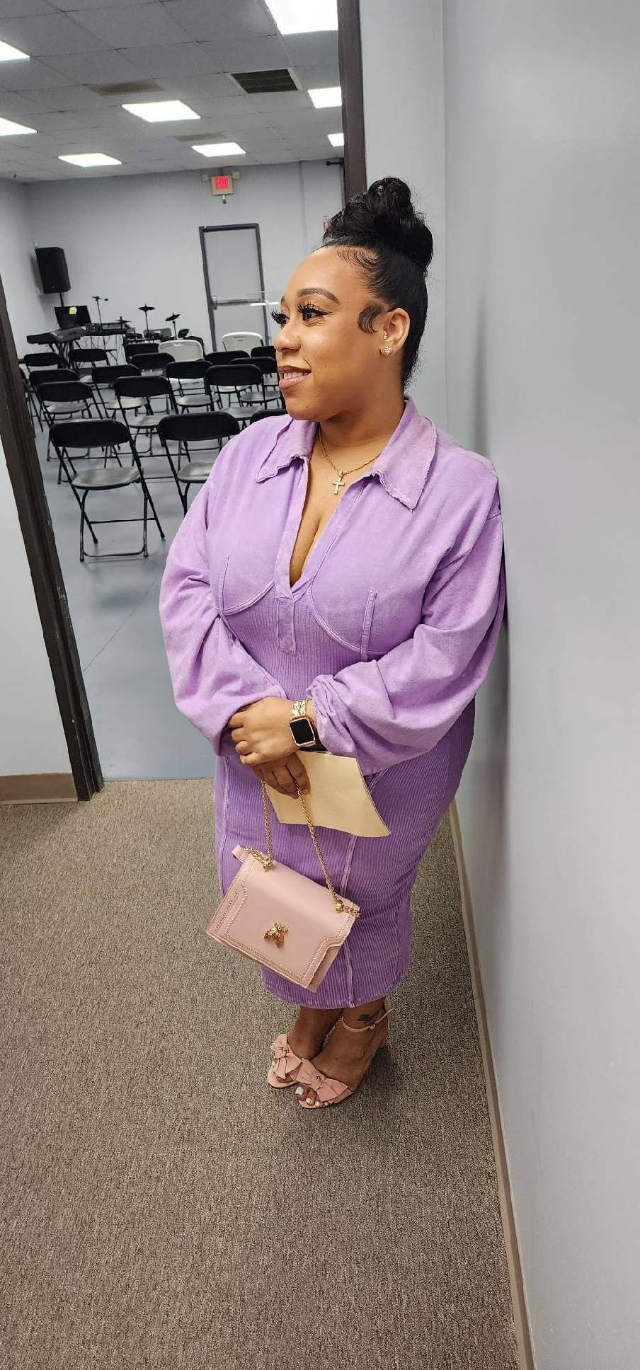 Woman in purple dress leans against wall, holding purse. Indoors, chairs and door visible.