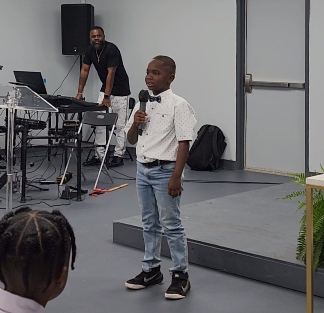 A young boy speaking into a microphone on a stage. A man plays music in the background.