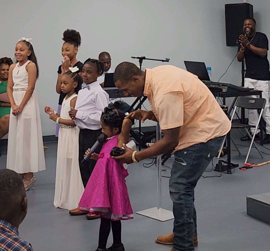 A man helps a young girl in a pink dress with a microphone, a choir stands behind them.