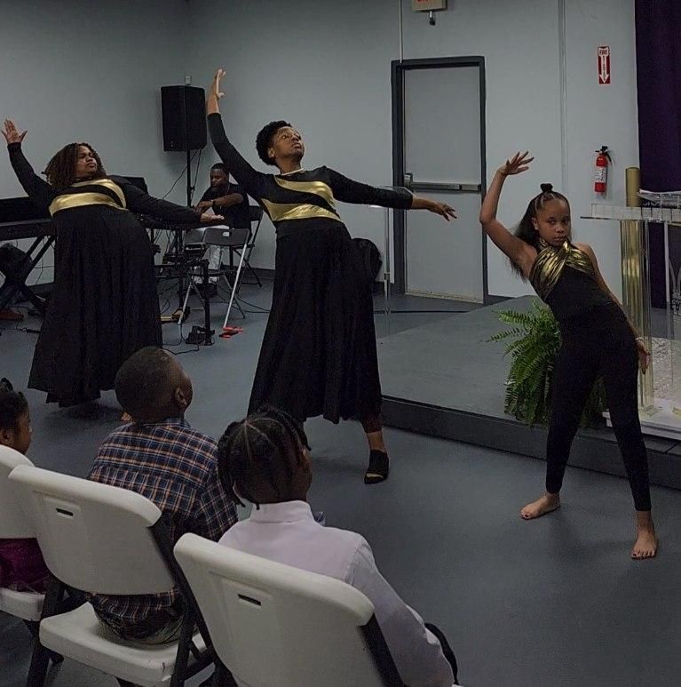 Dancers in black and gold perform in a room, observed by children. Church setting with band in the background.