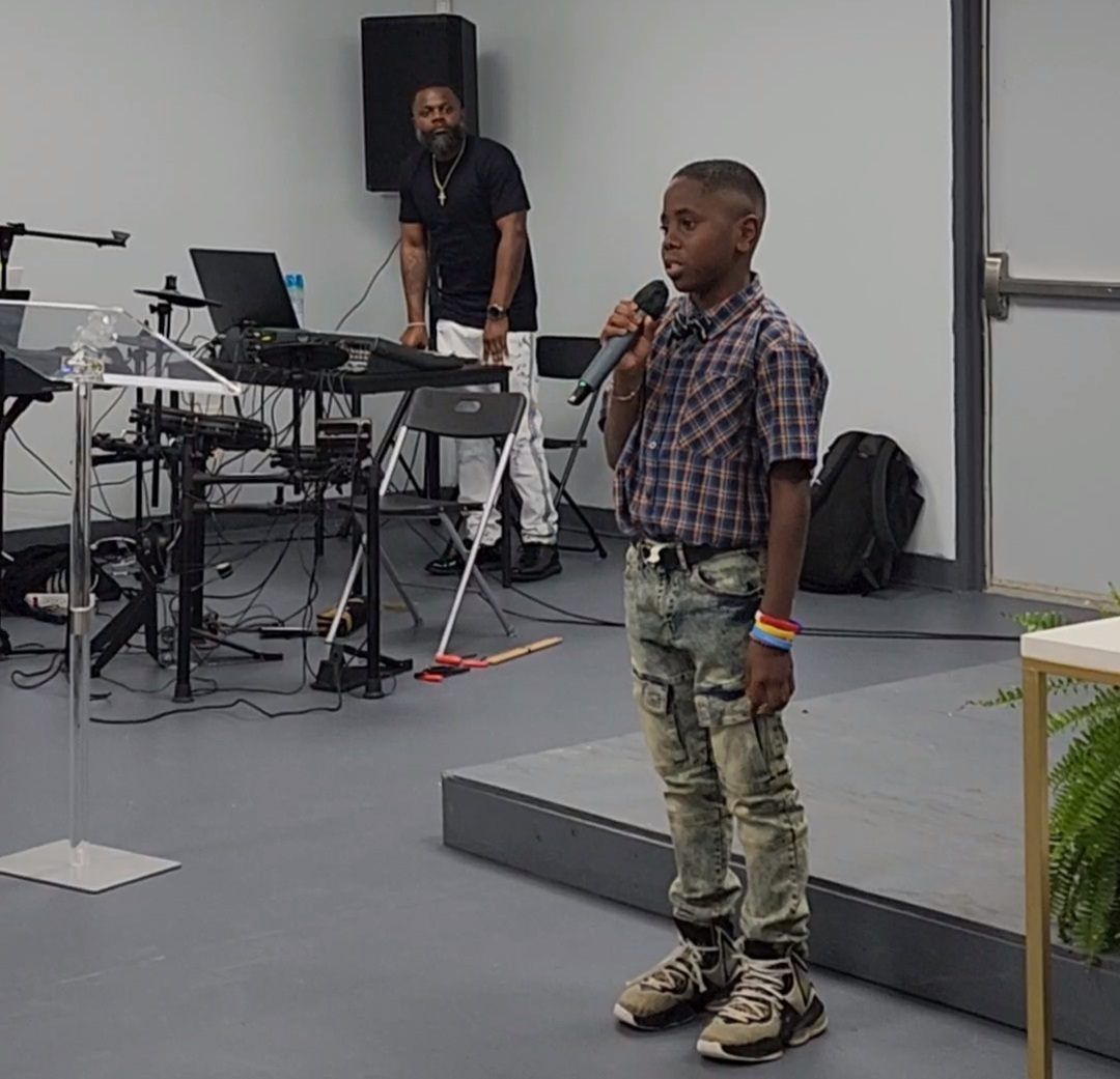 Young boy speaking into a microphone on a stage; man behind him at a keyboard; indoor setting.