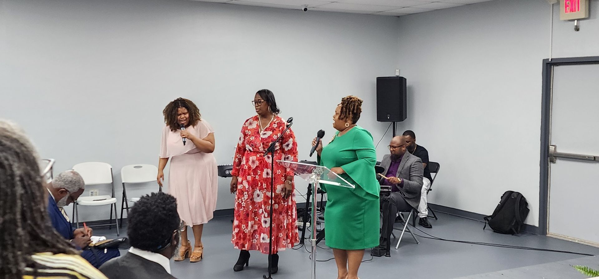 Three Black women stand at a podium and microphone in a room with other attendees.