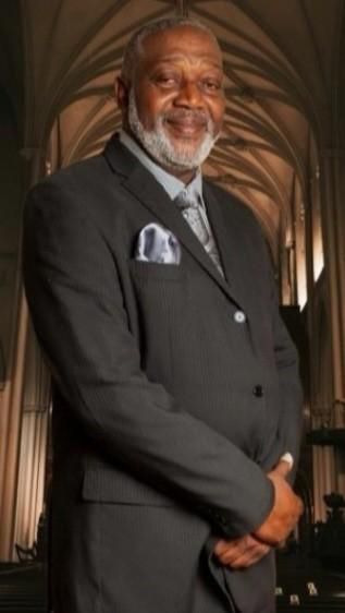 Man in a dark suit with a patterned tie, smiling, standing in a church-like setting.