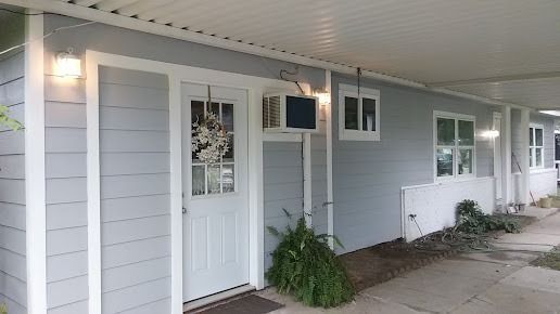 A light blue building with a white door, windows, and trim. A dark air conditioner is mounted above plants.