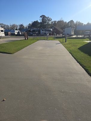 Concrete RV pad with water hookups, bordered by grass. Campers are visible in the background on a sunny day.