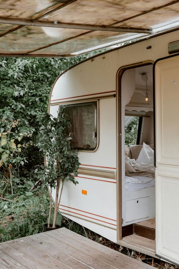 White vintage camper with an open door, parked next to lush greenery on a wooden deck.