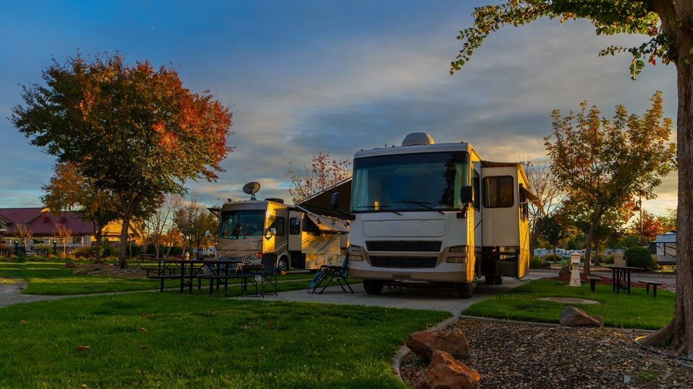 Campground with RVs parked on green grass; trees with fall foliage and sunset sky.