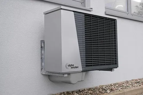 A silver Alpha Innotec heat pump unit mounted on a light gray exterior wall above a gravel ground surface.
