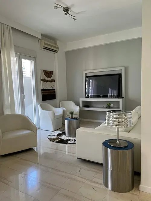 A modern living room with white armchairs, a white sofa, a metal side table with a lamp, and a wall-mounted television.