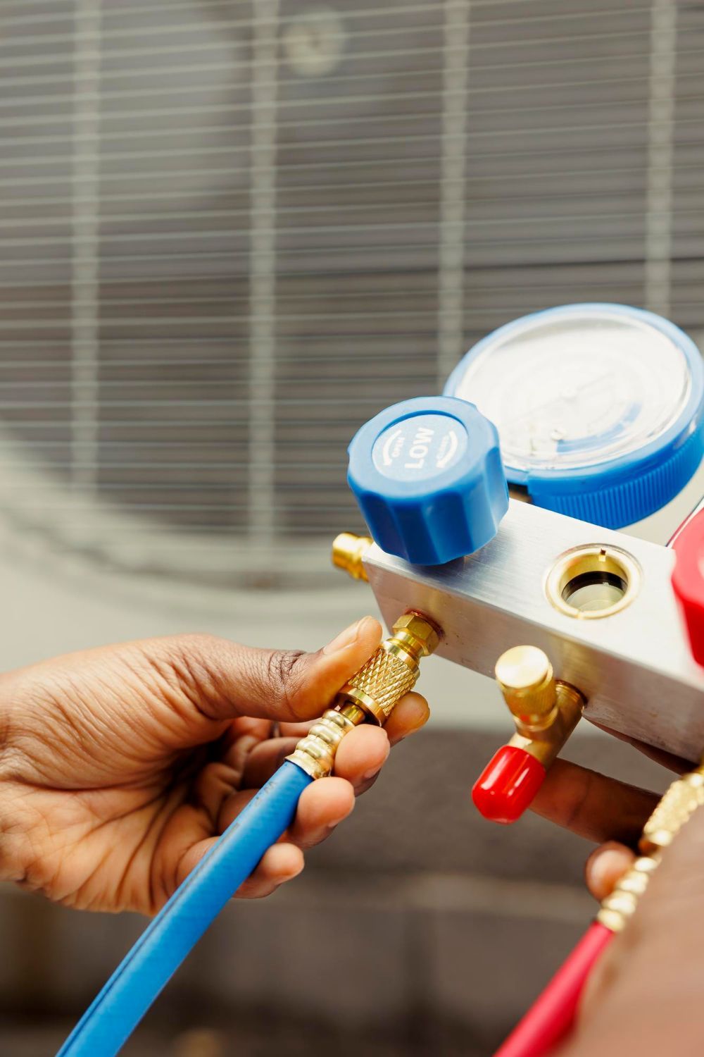 A technician's hands connect a blue hose to an HVAC manifold gauge set in front of an outdoor air conditioning unit.