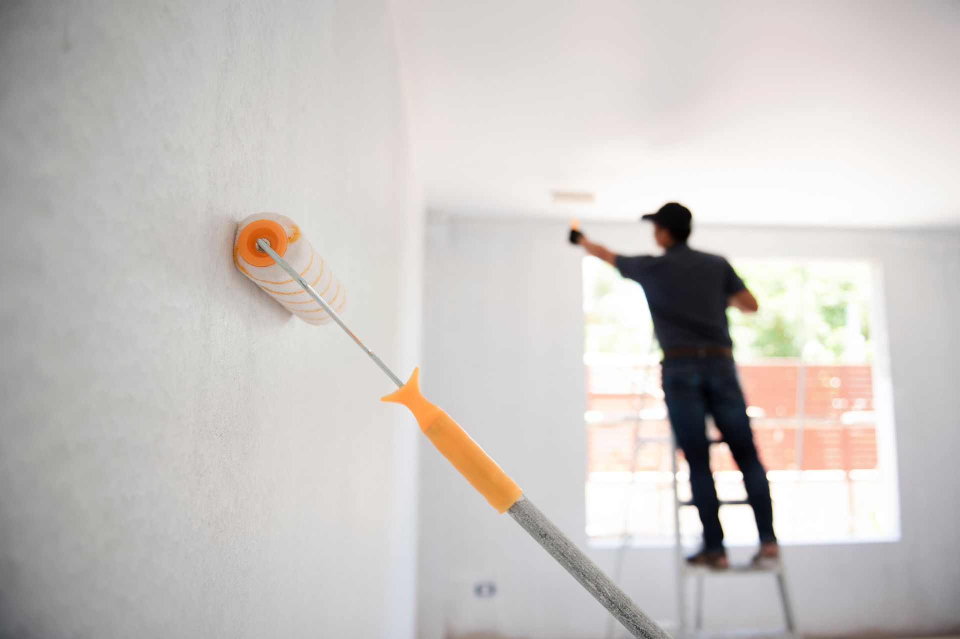 A person painting a white wall with a roller; standing on a stepladder in a bright room.