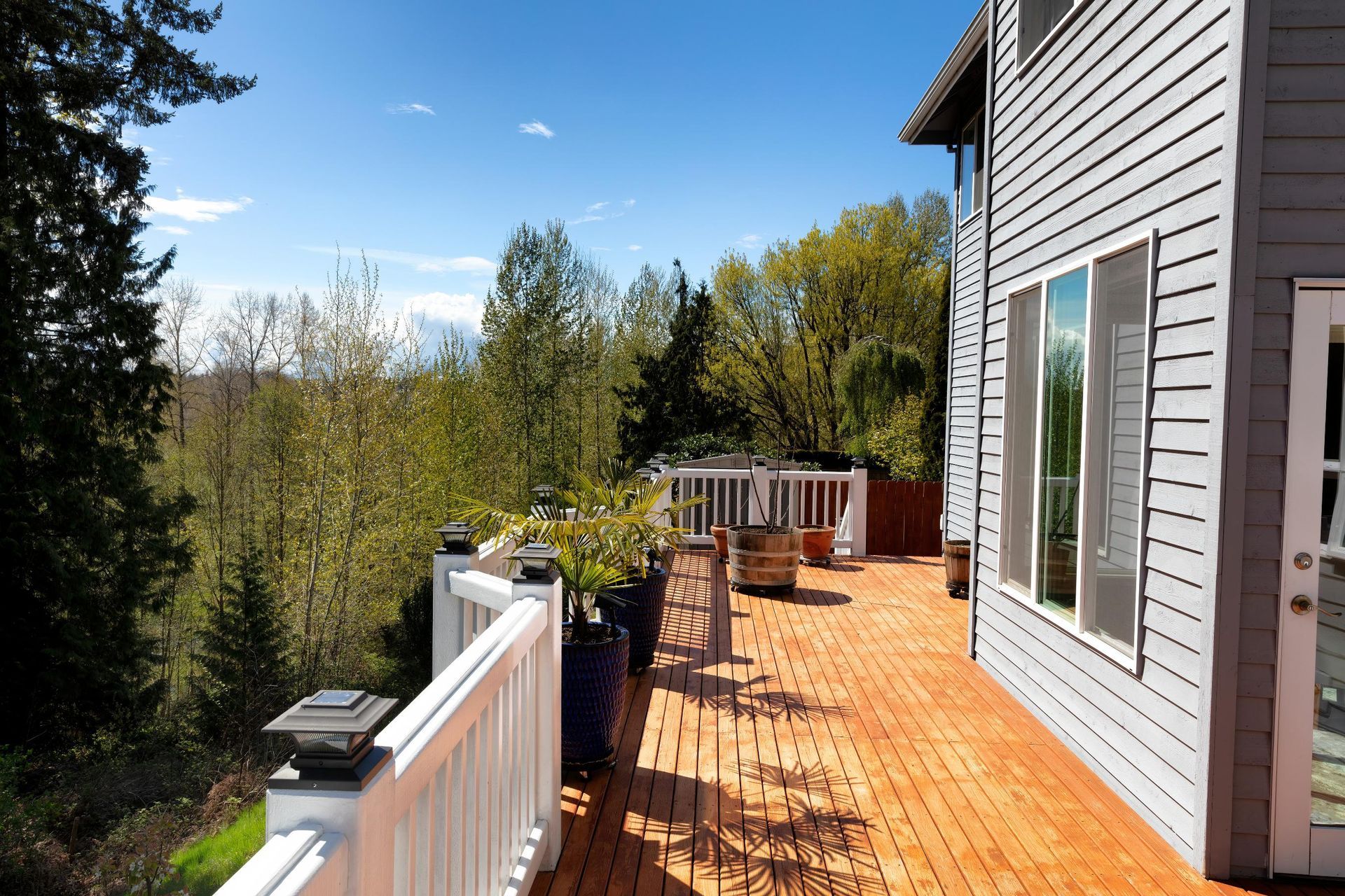Wooden deck with white railing beside a gray house, overlooking a lush green tree landscape under a blue sky.