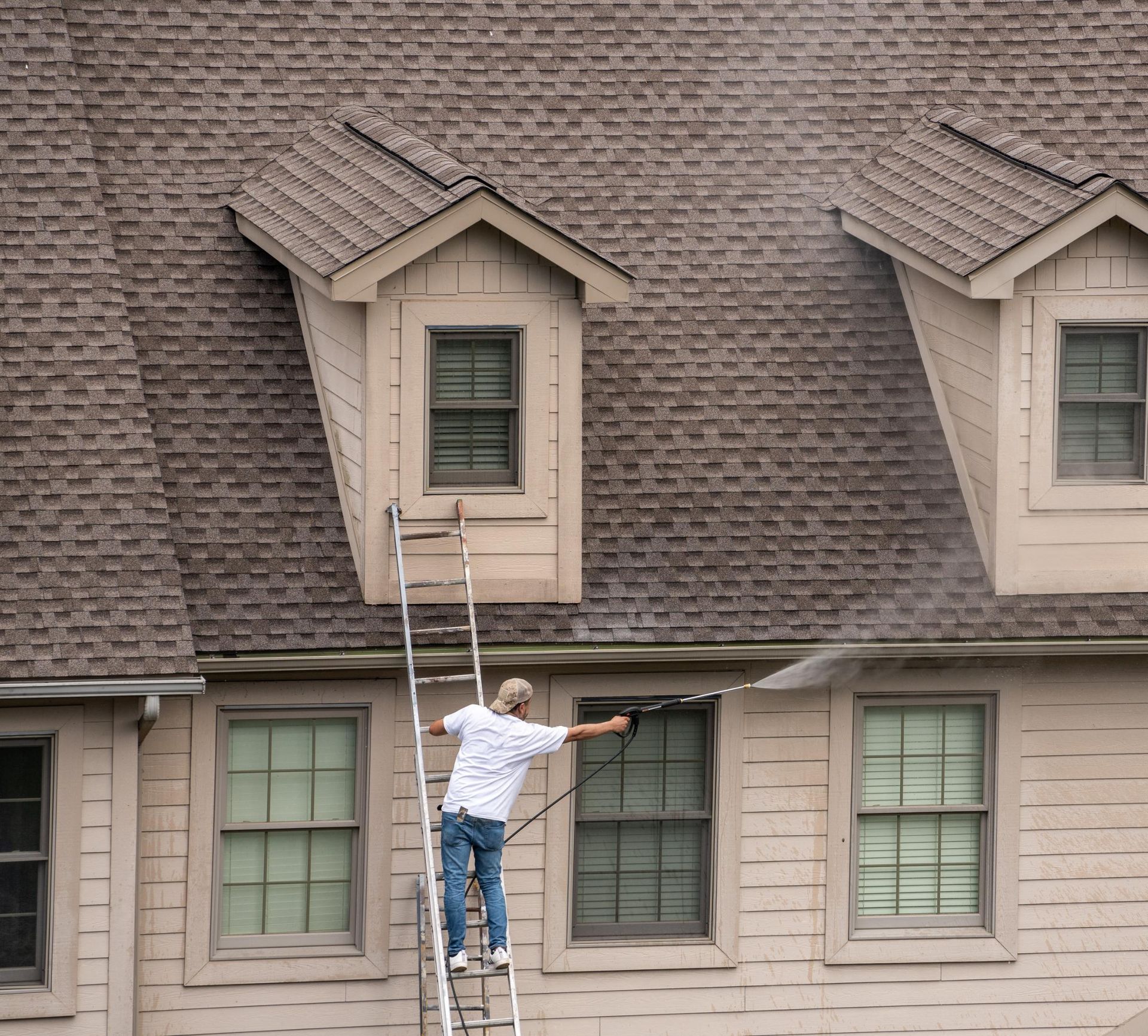 Man power washing the exterior of a house, standing on a ladder next to a window.