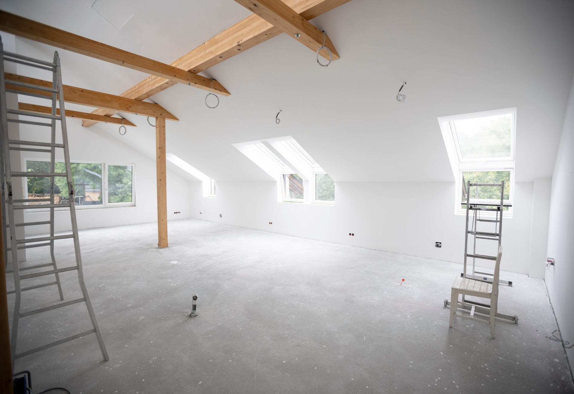 Spacious, white-walled attic room under construction, with wooden beams, ladders, and windows.