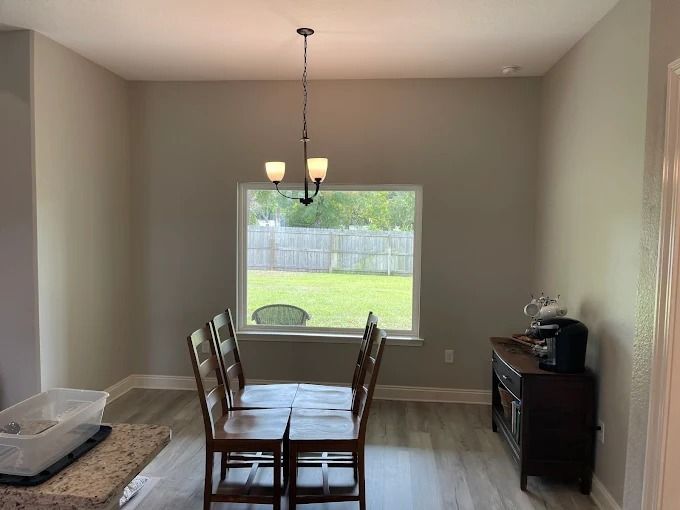Dining room with table, chairs, and a window overlooking a backyard. Gray walls, brown furniture, and light flooring.