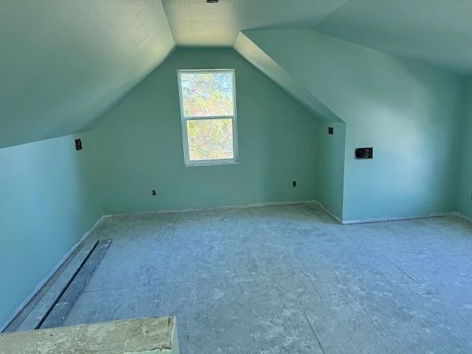 An empty, light-blue painted attic room with a window, exposed floor, and electrical outlets.